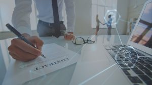 Man signing contract with right hand, glasses on the table near his left. Two people shaking hands upper right. A laptop computer lower right with a Bitcoin symbol superimposed