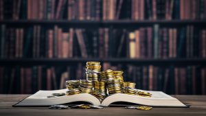 Stacks of coins sitting on top of an open book, on a table with a full bookshelf in the background