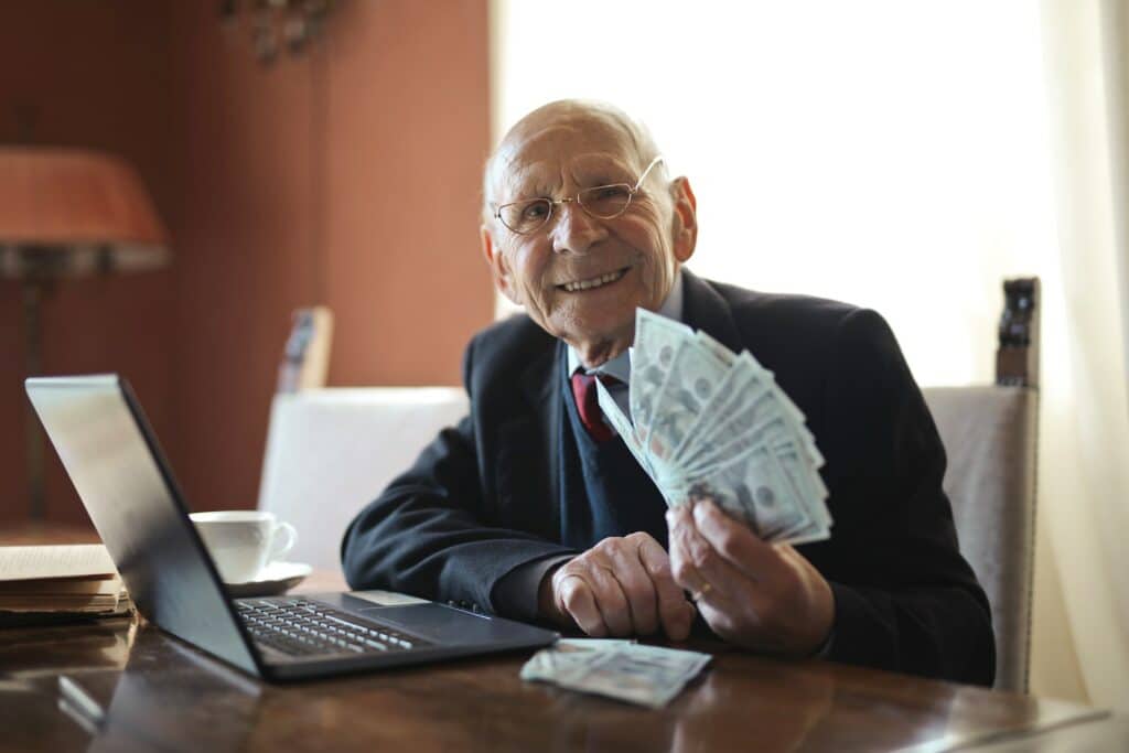 Old man in a suit with glasses, smiling as hands over a handful of money. Image courtesy of Andrea Piacquado at Pexels. 