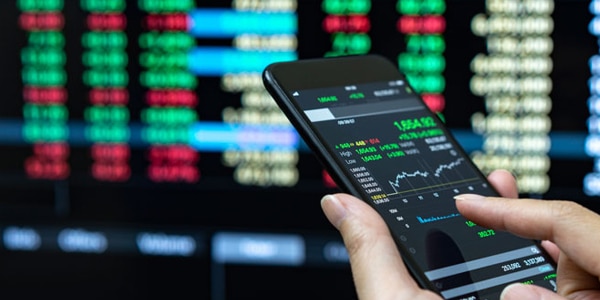 A cellphone held in a woman's left hand with a price chart. A quote board is in the background.