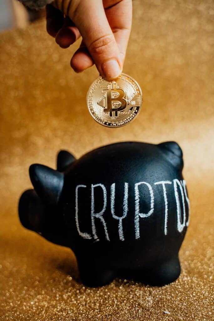 A hand depositing a Bitcoin into a black piggy bank with crypto's written on it. Image courtesy of Alexis Kozik on Pexels