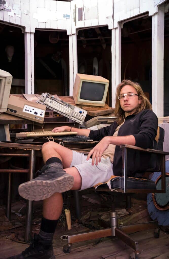 Young guy with taped glasses, seated nonchalantly and surrounded by vintage computer equipment. Photo courtesy of David Behar