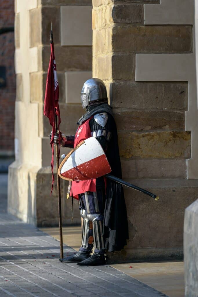 A knight in a full suit of armor with shield, sword, and flag outside of a building. Photo courtesy of the photoMIX company at Pexels