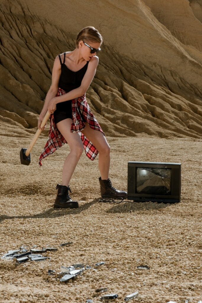 Young woman swinging a sledgehammer at a television sitting outside on some gravel. Image courtesy of cottonbro at Pexels. 